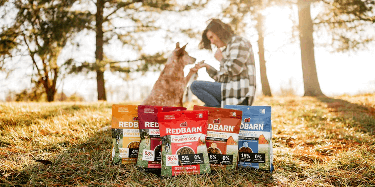 Woman sitting with a dog in a park, surrounded by various Redbarn pet food packages.